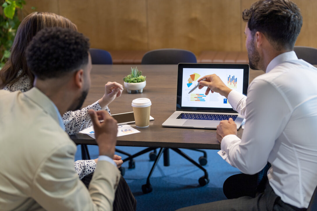 Person presenting to two others at a table, pointing to a computer screen.