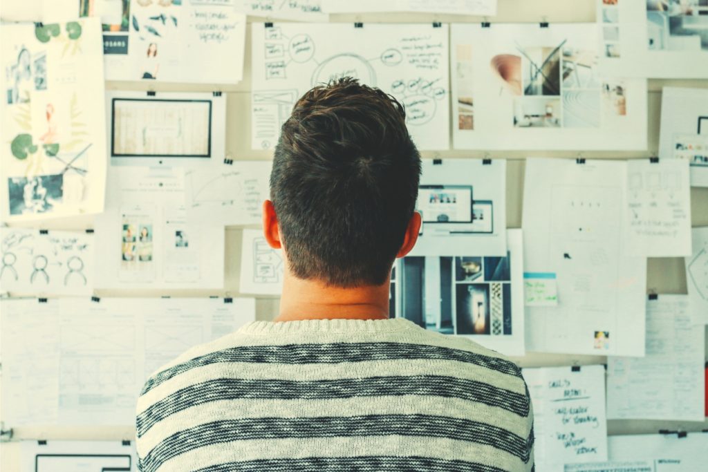 Image of a man looking at a bulletin board with many pieces of paper thumbtacked and linked together. 