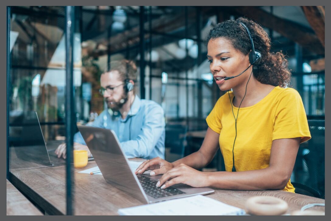 photo of two customer support employees sitting at a desk typing on their laptops and wearing headsets